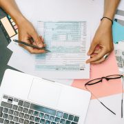 Pessoa preenchendo documentos financeiros diante de um laptop, com óculos e dinheiro sobre a mesa