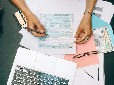 Pessoa preenchendo documentos financeiros diante de um laptop, com óculos e dinheiro sobre a mesa