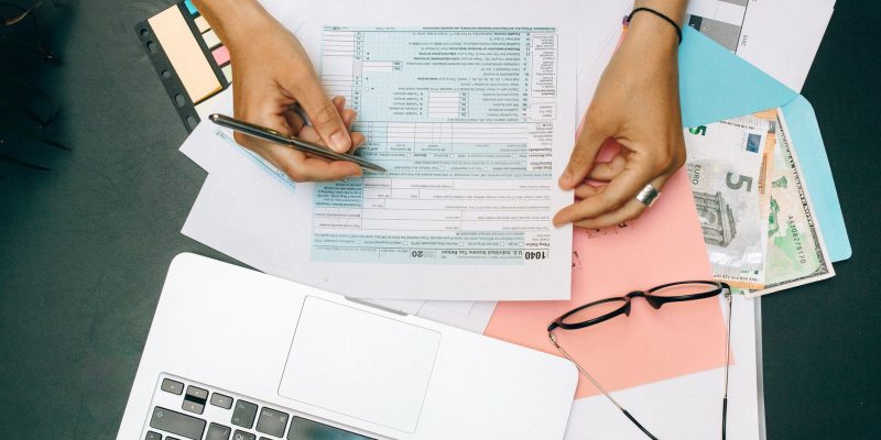 Pessoa preenchendo documentos financeiros diante de um laptop, com óculos e dinheiro sobre a mesa